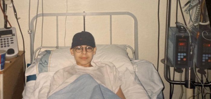 young boy with black baseball cap in hospital bed with white sheet and light blue blanket looking straight at the camera