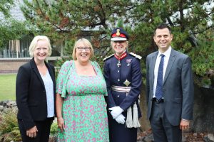 row of four people in a line for photo -  left to right, female blonde hair in navy suit, blonde hair and green and pink  dress, lord lieutenant Hilary wearing miltary like attire, man in navy suit