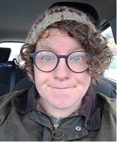 The person smiles at the camera. They have glasses and a hat and curly hair. 