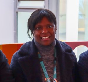 A woman with black hair is smiling at the camera. She wears a blue fluffy jacket and a blue lanyard.