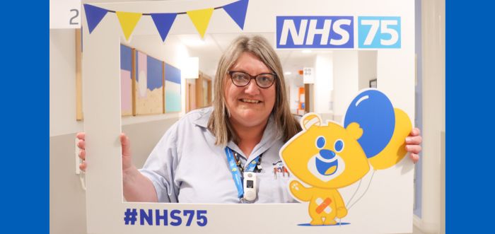 A woman is standing in a hospital corridor holding a white frame which says 'NHS 75' on it.