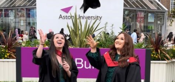 Two women in graduation robes throw their caps into the air in celebration infront of a sign reading Aston University.