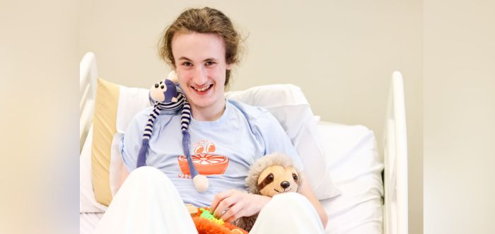 A teenage boy is sitting in a hospital bed. He has a blue and white striped monkey toy sitting on his left shoulder and a sloth toy on his knee.