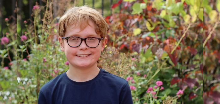 A young person is standing smiling in a garden. They have dark glasses and a blue navy long sleeved top on. There are lots of bright flowers and greenery behind them.