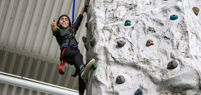 A child gives a thumbs up while holding onto an indoor climbing wall.