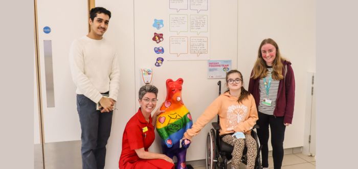 Three teenagers and a member of staff are visiting a hospital ward. They are next to a rainbow painted bear and a young person in a wheelchair is holding the bear's hand.