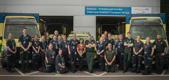 A group of Embrace team members are in their dark green and blue uniforms infront of the building and ambulances
