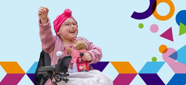 A young child in a wheelchair is raising one hand in the air and smiling widely.