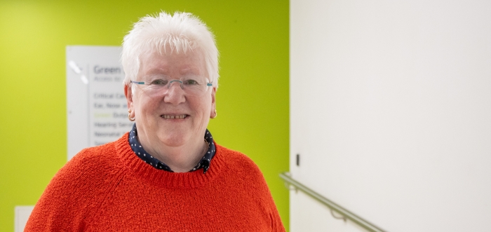 A woman with short white hair and a bright red jumper is standing in front of a bright green wall