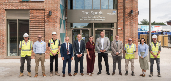 A line of adults stand in front of a new building. Some are wearing hi-vis. The sign on the building behind says "The Spark. Home of the National Centre for Child Health Technology"